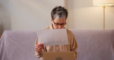 Latin kadın evdeki kanepede dizüstü bilgisayarla otururken istatistiksel verileri gözden geçiriyor. Kıdemli bayan, iş belgelerini konsantre bir ifadeyle analiz eder.