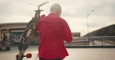 Aktif, mutlu, yaşlı, kırmızı tişörtlü kaykaycı gün batımından sonra şehir merkezinde kaykayla geziyor. Yaşlılar özgürlük ruhu kavramı. Yavaş çekim.