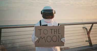 Uzay elbisesi içinde, üzerinde Ay 'a yazılmış karton posterlerle otostopçu bir kadın astronot. Kozmosun derinliklerini keşfeden İspanyol kadın kozmonot. Siber punk estetiği.