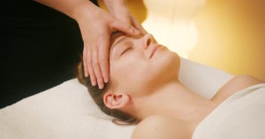 Spa Salonu 'nda yatakta uzanan ve yüz masajından zevk alan mutlu genç kadın. Lenfatik drenaj ve kaldırma efekti için kadın müşteri yüzüne masaj yapan kozmetik uzmanı. Güzellik, Cilt Bakımı.