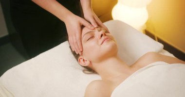 Spa Salonu 'nda yatakta uzanan ve yüz masajından zevk alan mutlu genç kadın. Lenfatik drenaj ve kaldırma efekti için kadın müşteri yüzüne masaj yapan kozmetik uzmanı. Güzellik, Cilt Bakımı.