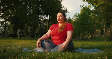 Vücut Portresi: Aşırı kilolu kadın Yoga yapıyor Sabahın köründe şehir dışında spor yapıyor, artı Yoga minderinin üzerinde oturan kadın boyutu.