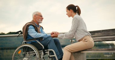 Genç bir kadın tekerlekli sandalyedeki yaşlı adamla el ele oturuyor. Yaya köprüsü üzerinde yürürken şefkatli destek bağlantısı ve sakin sohbet