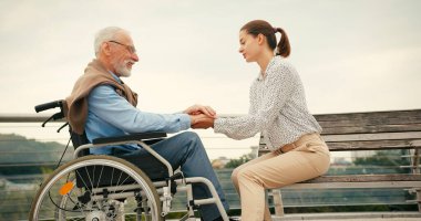Genç bir kadın tekerlekli sandalyedeki yaşlı adamla el ele oturuyor. Yaya köprüsü üzerinde yürürken şefkatli destek bağlantısı ve sakin sohbet