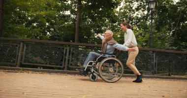 Parkta kadın tarafından kolları gerilmiş, tekerlekli sandalyedeki gülümseyen yaşlı adam. Nesiller arası arkadaşlık desteği ve mutluluk kavramı