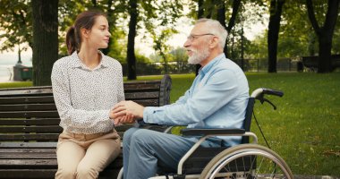 Tekerlekli sandalyedeki adam ve kadın el ele tutuşup parkta oynaşıyorlar. Sessiz bir bağ ve destek anı konfor ve arkadaşlığı iletir