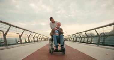 Genç bir kadın, yaşlı bir adamı bulutlu gökyüzünün altında köprüde itiyor. Huzurlu bir bağlantı anı ve birlikte yolculuğun kasıtlı temposu.