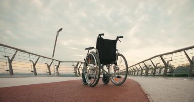 Günbatımında kaldırımlı köprüde tekerlekli sandalye boş duruyor. Sahne, ulaşılabilirlik ve hareket potansiyeli odaklı bekleme duygusunu uyandırır