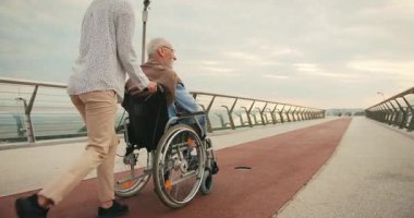 Genç kadın, yaşlı bir adamla tekerlekli sandalyede koşuyor ve kollarını uzatıp köprüde uçuyor. Birlik hissi ve paylaşılan sevinç güçlü bağlantıyı vurguluyor