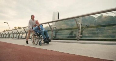 Genç kadın, yaşlı bir adamla tekerlekli sandalyede koşuyor ve kollarını uzatıp köprüde uçuyor. Birlik hissi ve paylaşılan sevinç güçlü bağlantıyı vurguluyor