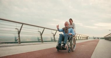 Genç kadın, yaşlı adamı tekerlekli sandalyede kollarını köprüye uzatarak şehir manzarasıyla itiyor. Neşeli ve aktif gezinti erişilebilirliği vurguluyor