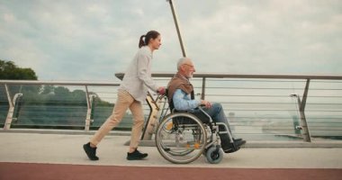 Genç kadın, yaşlı adamı tekerlekli sandalyede kollarını köprüye uzatarak şehir manzarasıyla itiyor. Neşeli ve aktif gezinti erişilebilirliği vurguluyor