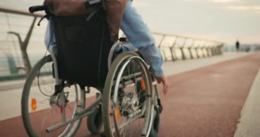 Tekerlekli sandalyeyi ileri itiyor. Köprü yolunda dönen tekerlekler hareket üzerine odaklanıyor. Bağımsız hareketlilik ve devam eden yolculuk kararlılığı önerir