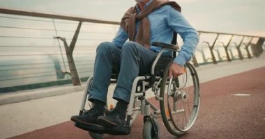 Tekerlekli sandalyeyi ileri itiyor. Köprü yolunda dönen tekerlekler hareket üzerine odaklanıyor. Bağımsız hareketlilik ve devam eden yolculuk kararlılığı önerir