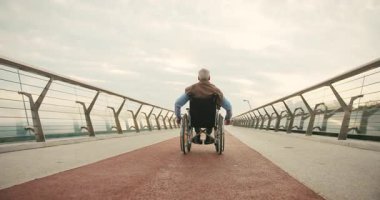 Tekerlekli sandalyedeki yaşlı adam, geniş, boş köprü geçidinde tekerlek çevirerek uzaklaşıyor. Bağımsız yolculuk yalnızlık ve kentsel ortam kavramına odaklanır.