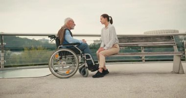 Genç bir kadın tekerlekli sandalyedeki yaşlı adamla el ele oturuyor. Yaya köprüsü üzerinde yürürken şefkatli destek bağlantısı ve sakin sohbet