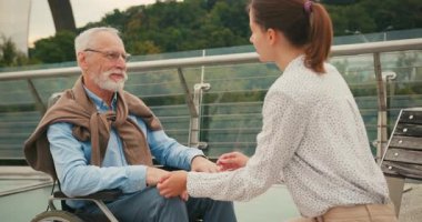 Genç bir kadın tekerlekli sandalyedeki yaşlı adamla el ele oturuyor. Yaya köprüsü üzerinde yürürken şefkatli destek bağlantısı ve sakin sohbet