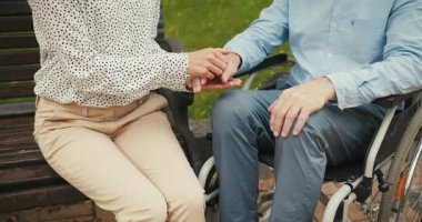 Tekerlekli sandalyedeki adam ve kadın el ele tutuşup parkta oynaşıyorlar. Sessiz bir bağ ve destek anı konfor ve arkadaşlığı iletir