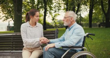 Genç kadın, parkta el ele tutuşan tekerlekli sandalyedeki yaşlı adamı destekliyor. Şefkatli bakım nesiller arası arkadaşlık ve duygusal destek
