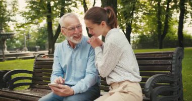 Kadın ve kıdemli adam parkta bankta oturup fotoğraf izlemek için akıllı telefon kullanıyorlar. Yürürken paylaşılan deneyim ve bağlantı anı