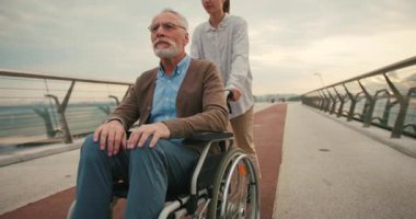 Tekerlekli sandalyedeki adam, eli nazikçe omzuna yerleştirilmiş bir kadın tarafından destekleniyor. Güçlü rahatlık ve destek duygusu derin birlik ve bağlantıyı vurguluyor