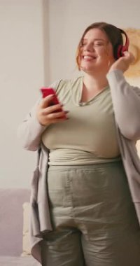 Ayrıca kulaklıklı cüsseli bir kadın elinde akıllı telefonuyla müzik dinliyor. Gülümseyen kadın hafifçe dans ediyor omuzlarını şarkıya doğru hareket ettiriyor evde mutlu görünüyor.
