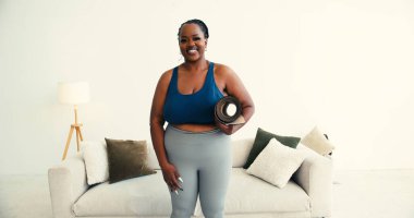 Kendine güveni tam, Afrikalı Amerikalı kadın, yoga minderini katlamış. Vücut pozitifliğini somutlaştıran kişi ev konforunda sağlıklı yaşam tarzını benimser.