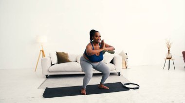 Artı boyut siyah kadın dizlerini büker vücudu derin çömelme pozuna sokar. Eller birbirine bastırarak dengeyi yaratırken kaslar güçlü bir hareketle geriliyor