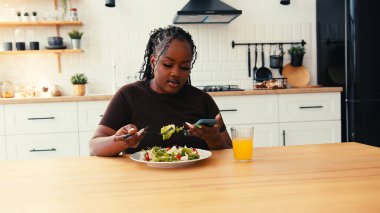 Mutfak masasında oturan şişman siyah kadın taze salata yiyor. Kadın telefona bakar sağlıklı yemek yer ve çevrimiçi dünyaya bağlanır.