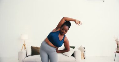 Kendine güveni tam, spor kıyafetli zenci bir kadın modern oturma odasında kol esnetme hareketleri yapıyor. Kadın, vücut pozitifliğini ve sağlıklı yaşam tarzını benimser.