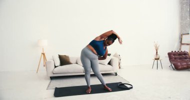 Kendine güveni tam, spor kıyafetli zenci bir kadın modern oturma odasında kol esnetme hareketleri yapıyor. Kadın, vücut pozitifliğini ve sağlıklı yaşam tarzını benimser.