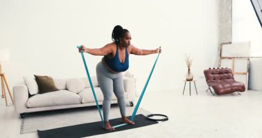 Artı boyut siyah kadın oturma odasında direnç bandı kullanarak egzersiz yapıyor. Dişi kas geliştirmeye ve kilo vermeye odaklanıyor