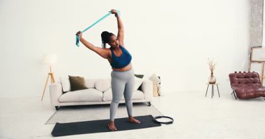 Ayrıca evde lastik bantla egzersiz yapan Afrikalı Amerikalı bir kadın. Kadın esneme bandını farklı yönlere doğru esnetir.