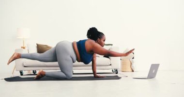 Aşırı kilolu Afrikalı Amerikalı kadın paspasın üzerinde sağ kolu ve karşı bacağı uzatarak dengede duruyor. Gövde dizüstü bilgisayarın önünde video öğreticisi ile hizalanır