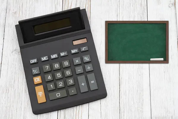在风吹日晒的木桌上装有黑板的黑色计算器的教育费用 — 图库照片