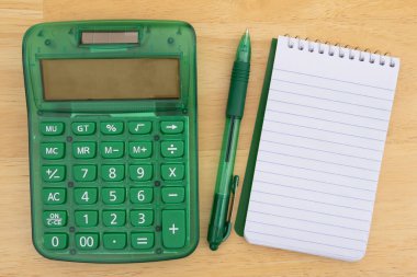 Tahta bir masanın üzerine yerleştirilmiş yeşil hesap makinesi, kalem ve defter. Mali, muhasebe ve bütçe kavramı.