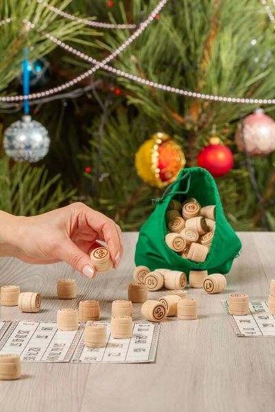 Woman\'s hand holding a barrel for a game in lotto. Wooden lotto barrels with bag and game cards, Christmas fir tree with toy balls and garlands on the background. Board game lotto.