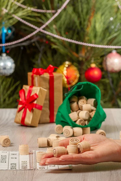 Woman\'s hand holding barrels for a game in lotto. Wooden lotto barrels with bag and game cards, Gift boxes, Christmas fir tree with toy balls and garlands on the background. Board game lotto.