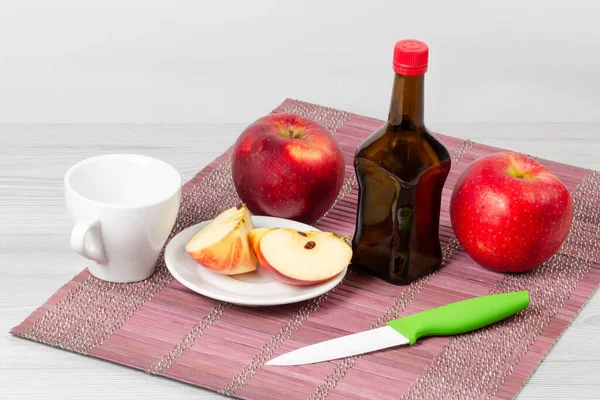Apple vinegar in a glass bottle with a cup and fresh red apples with a knife on a bamboo napkin. View from above.