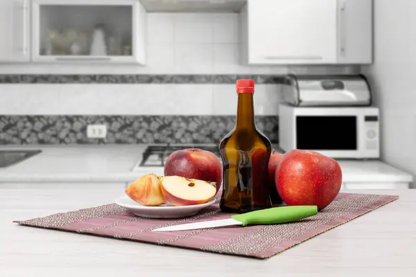 Apple vinegar in a glass bottle and fresh red apples with a knife on a bamboo napkin in the kitchen.