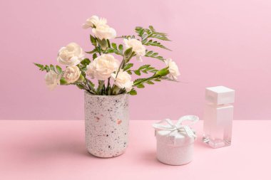Vazoda bir buket beyaz karanfil, hediye kutusu ve pembe arka planda bir şişe parfüm..