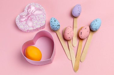 Ahşap kaşıklarda renkli Paskalya yumurtaları ve pembe arka planda bir hediye kutusu. Üst görünüm.