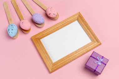 Ahşap kaşıklarda renkli Paskalya yumurtaları, fotoğraf çerçevesi ve pembe arka planda hediye kutusu. Üst görünüm.