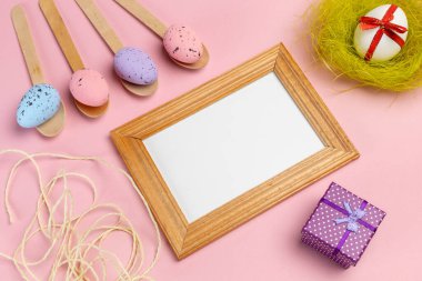 Ahşap kaşıklarda renkli Paskalya yumurtaları, fotoğraf çerçevesi, bir yuva ve pembe arka planda bir hediye kutusu. Üst görünüm.
