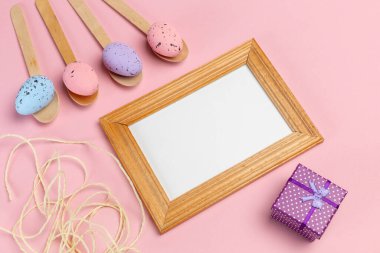 Ahşap kaşıklarda renkli Paskalya yumurtaları, fotoğraf çerçevesi ve pembe arka planda hediye kutusu. Üst görünüm.