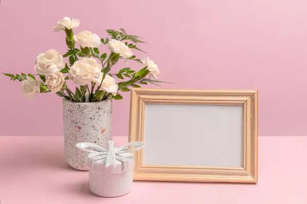 Bouquet of white carnations in a vase, a gift box and a photo frame with copy space on the pink background.