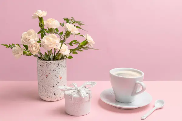 Bouquet of white carnations in a vase, a gift box and a cup of coffee on the pink background.
