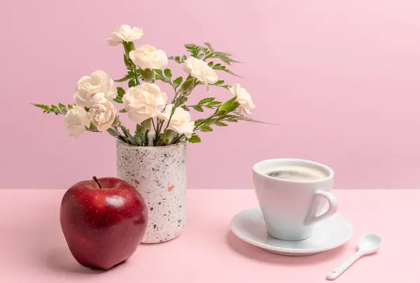 Bouquet of white carnations in a vase, a red apple and a cup of coffee on the pink background.