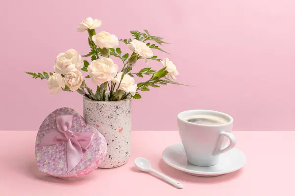 Bouquet of white carnations in a vase, a gift box and a cup of coffee with a spoon on the pink background.