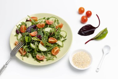 Taze domatesli salata, salatalık, roka, fesleğen, çatallı ıspanak. Susamlı kase ve bir kaşık. Beyaz arka plan. Sağlıklı sebze diyeti menüsü. Üst görünüm.